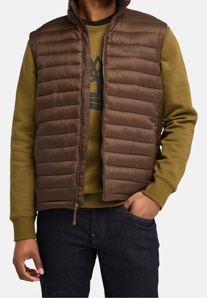 Homme portant un gilet matelassé marron sur un sweat-shirt vert olive et un jean foncé, avec une main dans la poche et le gilet partiellement zippé.