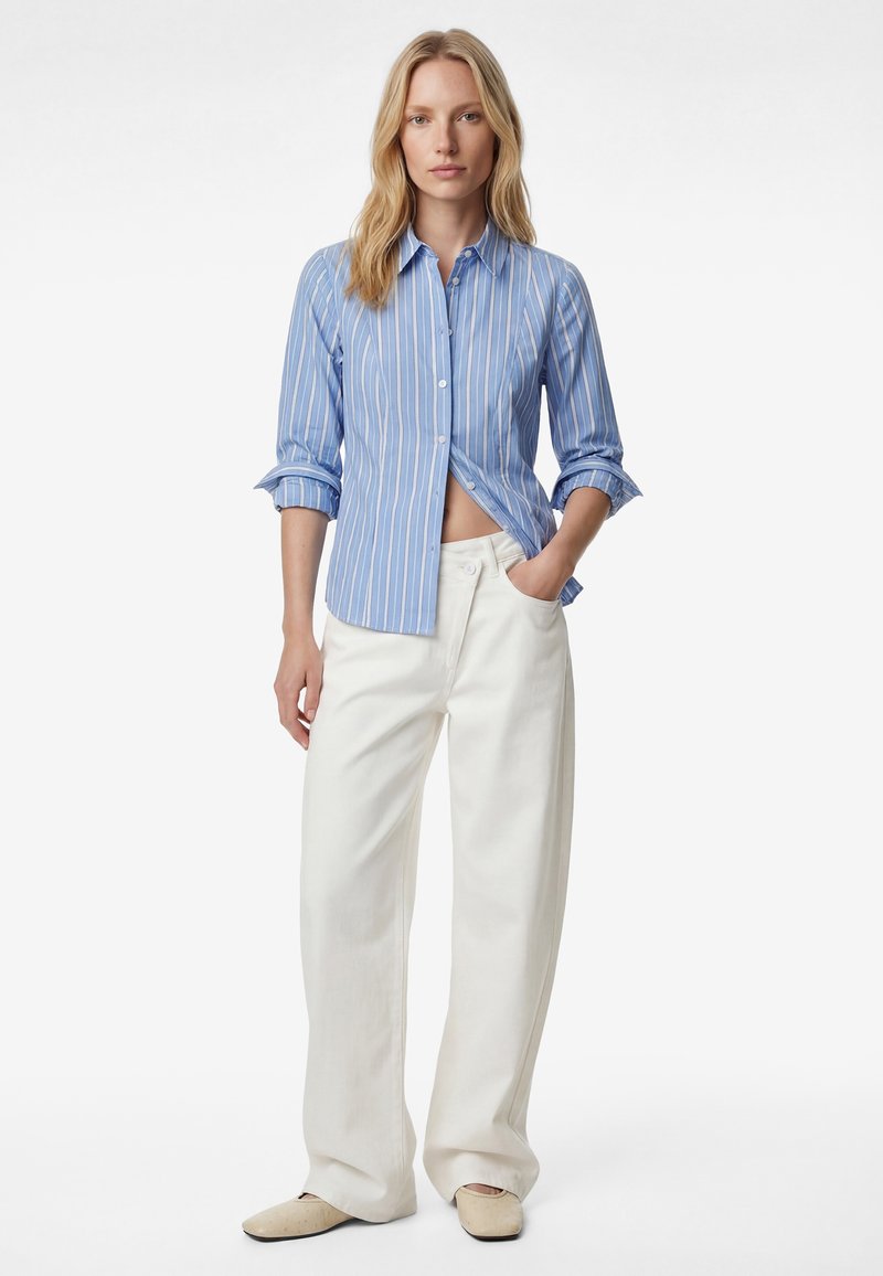 Kvinde stående iført en blåstribet skjorte med knapper og opkørte ærmer, hvide bukser med vide ben og beige sko, med hånden i lommen.