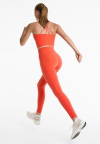 Fényes narancssárga sportos crop top és leggings, sima textúrával. A toppon vékony keresztbe fonódó pántok találhatók, aktív öltözködéshez tervezve.