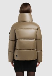 Chaqueta de plumas color caqui con un alto cuello, diseño acolchado y textura brillante. Presenta una pequeña etiqueta de logo negra en la esquina inferior izquierda.