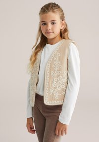 Gilet beige ricamato con motivi floreali bianchi, con apertura frontale, indossato sopra una camicia bianca e pantaloni a coste marroni, caratterizzato da cuciture intricate.