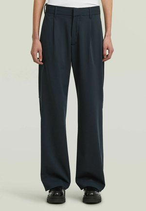 Pantaloni neri larghi con passanti per cintura, pieghe frontali e scarpe nere robuste indossati da una persona in piedi contro uno sfondo semplice.