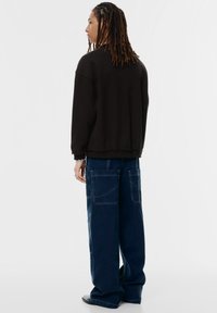 Hosszú raszta hajjal rendelkező személy, fekete, nagyméretű pulcsit és széles szárú, sötétkék nadrágot visel, amelyen nagy hátsó zsebek találhatók, oldalra áll.