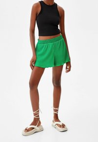 Zelené skládací šortky s elastickým pasem a rozšířeným střihem, spárované s přiléhavým černým topem a béžovými sandály na pásky.