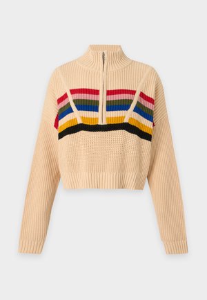 Personne portant un pull en maille beige avec des rayures horizontales colorées, un jean large bleu clair usé et des tongs noires, debout sur un fond blanc.
