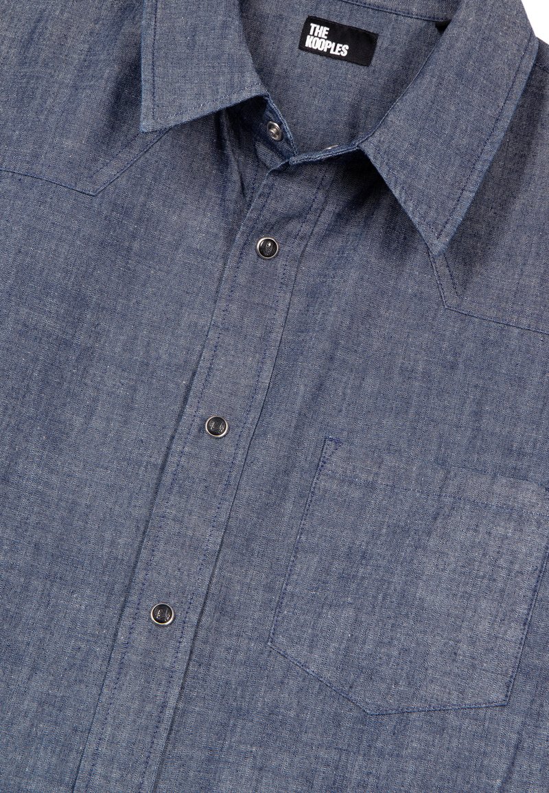 Denimskjorta med knappar i mörkblått, med spetsig krage, bröstficka och metallknappar med strukturerad yta.