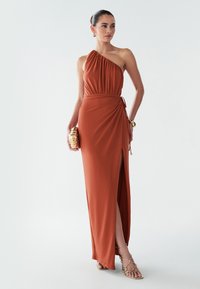 Šaty v cihlové barvě, s jedním ramínkem, s nařaseným materiálem, bočním rozparkem a zavázaným detailem v pase. Doplněné zlatou kabelkou a sandály na podpatku.