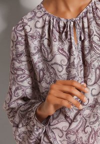 Helllila Bluse mit Paisley-Muster, gerafftem Ausschnitt und Schleifendetail. Lange Ärmel mit Knopfmanschetten. Weicher, fließender Stoff.
