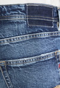 Primer plano de unos jeans de mezclilla azul oscuro que muestra la textura de la tela, costuras amarillas, bolsillos traseros y una pequeña etiqueta roja en la parte inferior derecha.