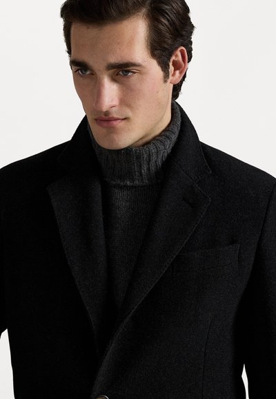 Black wool coat with a tailored silhouette and notched lapels, worn over a dark gray ribbed turtleneck sweater, highlighting textures and layering.