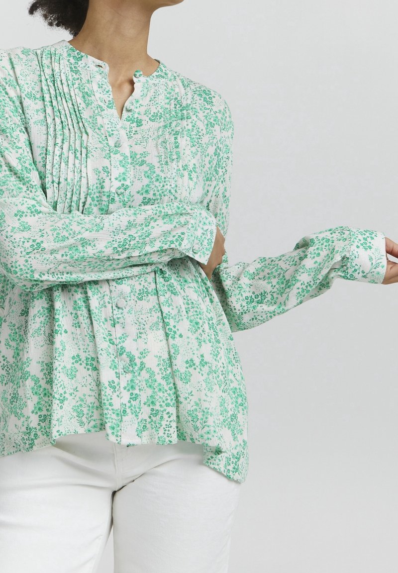 Long-sleeve blouse in white with green floral print, featuring pleats, a buttoned neck, and a slightly flared hem for a relaxed fit.