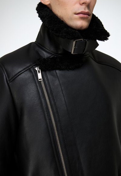 Schwarze Lederjacke mit hohem Kragen und schwarzem Lammfellfutter, seitlichem Reißverschlussdetail und matter Oberfläche.
