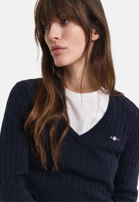 Navy-blaues Kabelstrick-Pullover mit V-Ausschnitt, über einem weißen Hemd getragen. Besitzt ein kleines Logo auf der Brust. Weiche Textur, gerippte Bündchen.