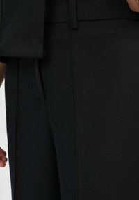 Gros plan sur une personne portant un pantalon noir sur mesure avec une poche latérale, une main reposant près de la taille, et une partie d'une veste noire assortie.