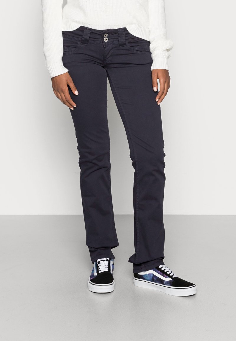 Osoba v tmavých slim-fit kalhotách, bílém svetru a černých teniskách s galaktickým vzorem po stranách, stojící na šedé podlaze.