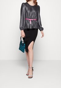 Metallisch graues Longsleeve-Oberteil mit gepufften Schultern, pinkem Gürtel, schwarzem Ripprock mit seitlichem Schlitz und tealfarbener gewebter Handtasche. Schwarze Riemchenheels.