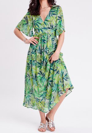 Femme en robe midi verte à fleurs avec manches volantes, col en V et sandales argentées, debout sur fond blanc.