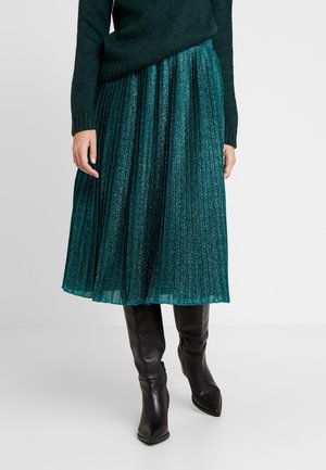 Metaliczna, plisowana spódnica midi w kolorze morskim, zestawiona z czarnymi, skórzanymi kozakami za kolano o spiczastym nosku oraz ciemnozielonym swetrem dzianinowym.
