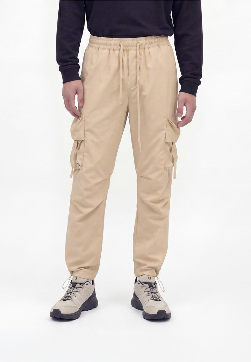 Personne portant un pantalon cargo beige avec taille élastique et poches latérales, associé à des chaussures de randonnée béiges et noires et un haut à manches longues noir.