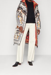 Abrigo acolchado con intrincados patrones florales en negro, naranja y blanco; forro interior rojo. Combinado con un suéter negro, pantalones blancos y botas negras.