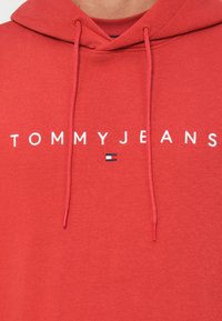 Červená mikina s kapucňou vyrobená z mäkkého materiálu, s bielym nápisom "TOMMY JEANS" a malým modro-bielo-červeným logom. Šnúrky s rovnakým zakončením.