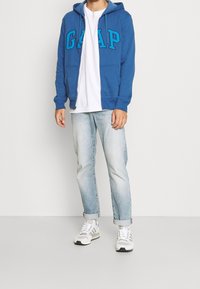 Homme portant un sweat à capuche bleu GAP, un t-shirt blanc, un jean bleu clair retroussé et des baskets blanches Adidas, debout devant un fond blanc uni.