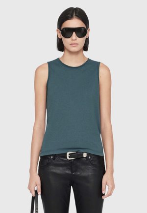 Woman with short dark hair wearing large black sunglasses, a sleeveless dark teal top, black leather pants, and holding a black strap.