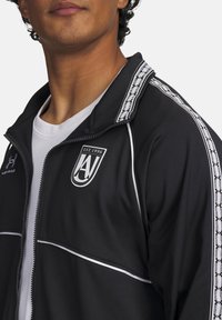 Black athletic zip-up jacket with white accents, featuring a logo and patterned trim on the shoulders. Made from a smooth fabric.