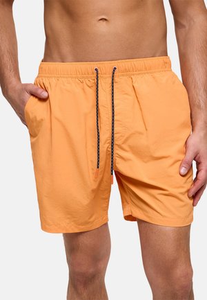 Hombre vistiendo pantalones cortos de baño naranjas con cordones negros y bolsillos laterales, de pie con las manos cerca de los bolsillos contra un fondo liso.