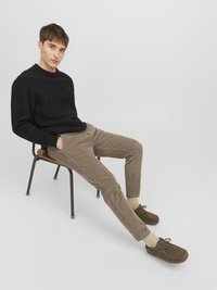 Maglione a maglia nera, pantaloni beige in corduroy e scarpe in suede marroni. Seduto su una sedia di metallo nera con sedile in legno. Sfondo neutro.