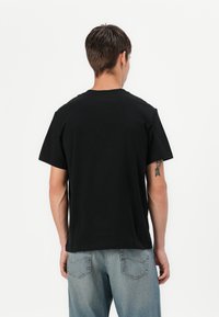 Tricou negru din bumbac, cu guler rotund și mâneci scurte, purtat de o persoană care poartă blugi din denim deschis la culoare. Design simplu, fără decor.