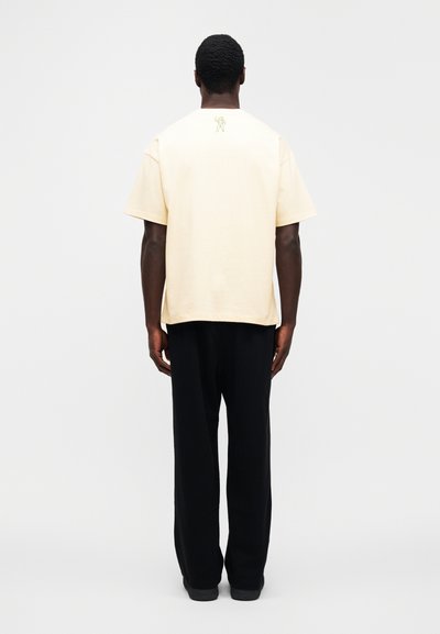 Μπεζ βαμβακερό t-shirt με κο ντά μανίκια, που έχει ένα διακριτικό πράσινο γραφικό στην πλάτη. Συνδυάζεται με φαρδιά μαύρη παντελόνια.