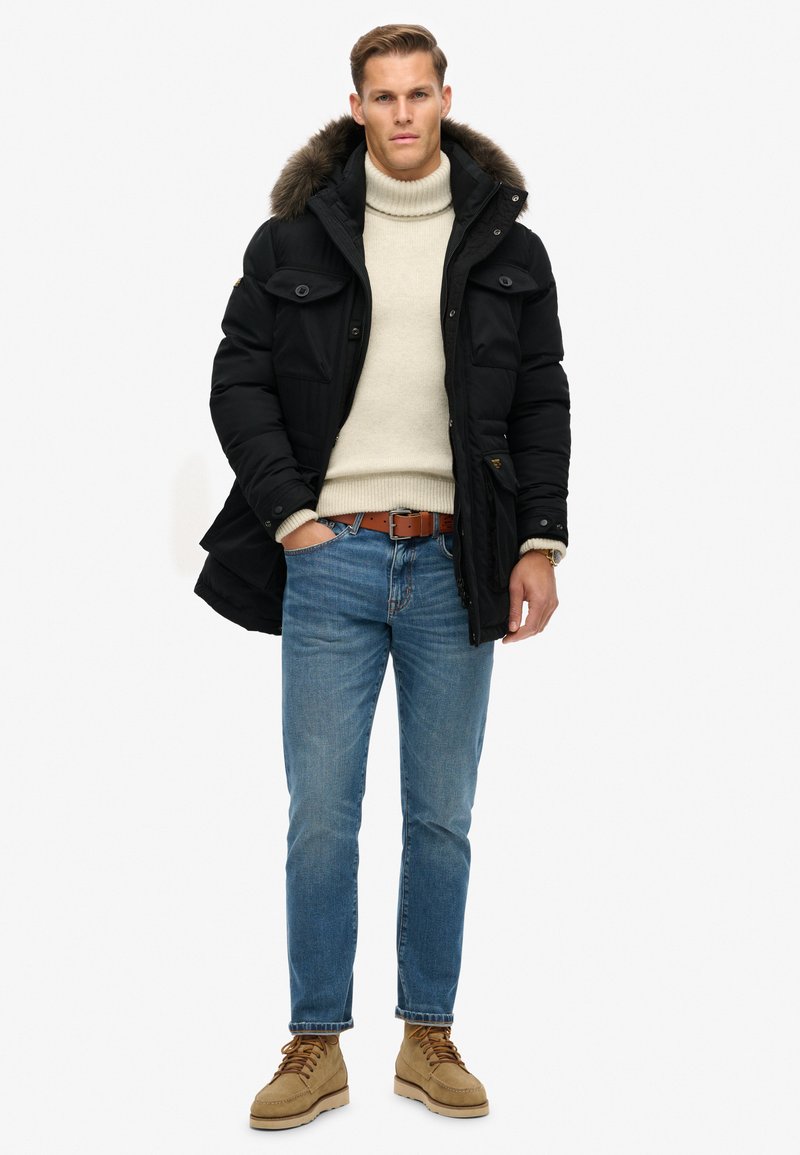 Man in zwarte parka met bontkap, crèmekleurige turtleneck trui, blauwe jeans, bruine riem en tan kleurige veterschoenen, staand met één hand in zijn zak.