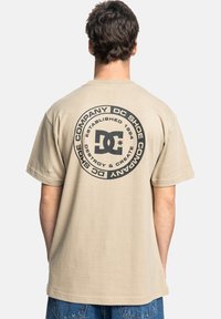 Béžové bavlněné tričko s velkým kulatým černým grafikou s nápisem "DC Shoe Company" a "Established 1994" na zadní straně. Krátké rukávy, volný střih.