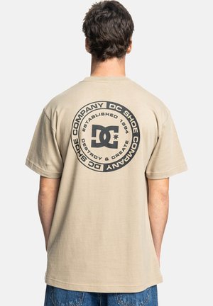Bežové bavlnené tričko s veľkým okrúhlym čiernym grafickým motívom na zadnej strane, ktorý obsahuje nápisy "DC Shoe Company" a "Established 1994". Krátke rukávy, voľný strih.