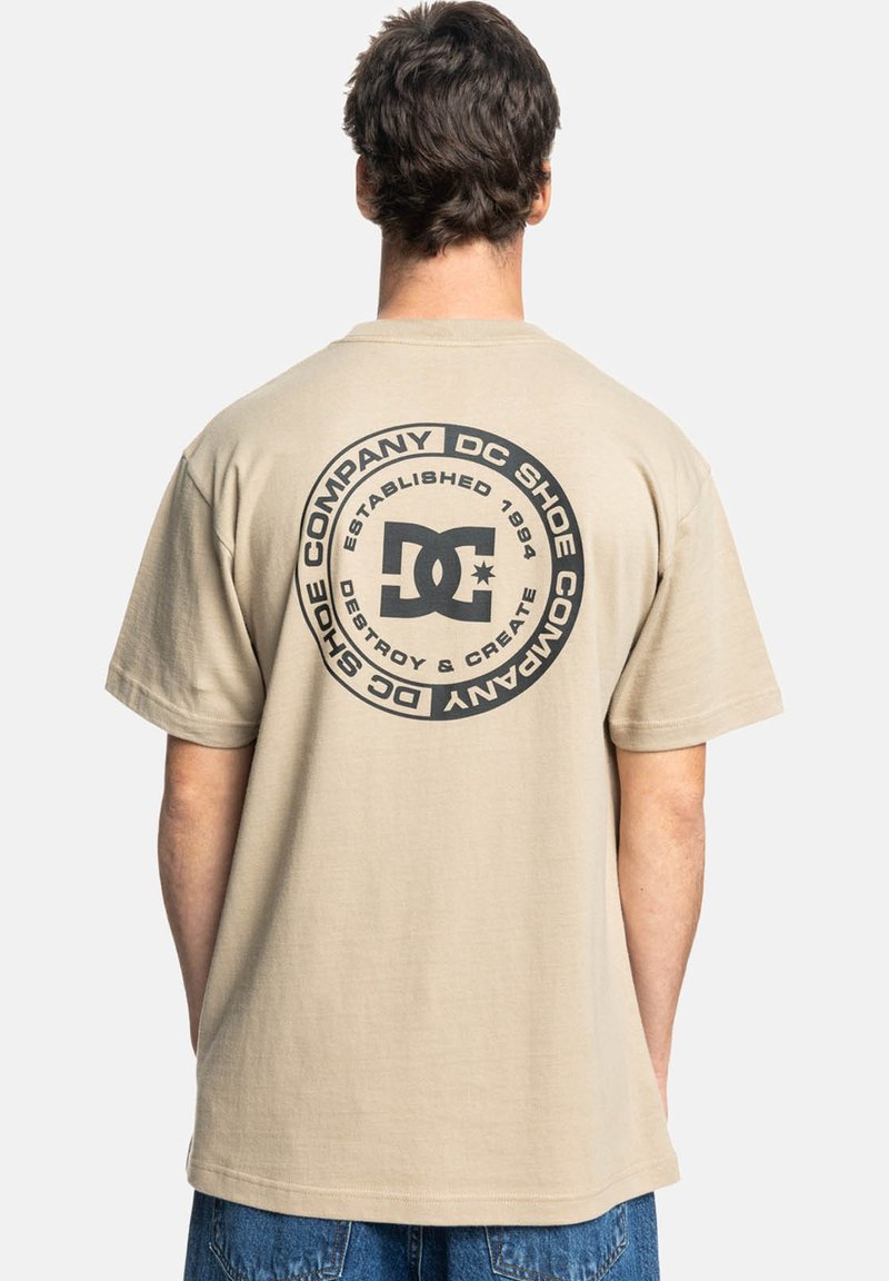 Béžové bavlněné tričko s velkým kulatým černým grafikou s nápisem "DC Shoe Company" a "Established 1994" na zadní straně. Krátké rukávy, volný střih.