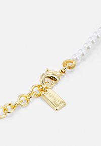 Gold metal chain with a lobster clasp, featuring white pearls, and a rectangular tag engraved with "COACH." Smooth texture and polished finish.