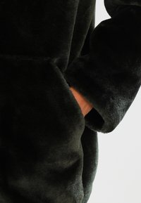 Tmavě zelený fleecový oděv s měkkou texturou, který má přední kapsu a volné rukávy. Tkanina vypadá nadýchaně a útulně.