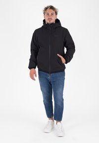 Zwarte capuchonjack met een ritssluiting aan de voorkant, elastische manchetten en zijzakken, gecombineerd met blauwe denim jeans en witte sneakers.