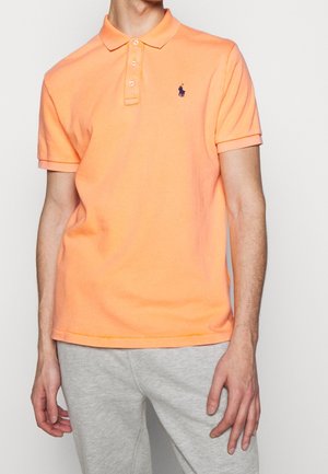 Pomarańczowa koszulka polo z kołnierzykiem, trzema guzikami i krótkimi rękawami, z małym niebieskim logo na piersi. Noszona z szarymi spodniami dresowymi.