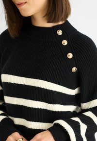 Black and white striped knitted sweater with a high neckline, ribbed texture, and gold button accents on the shoulder.