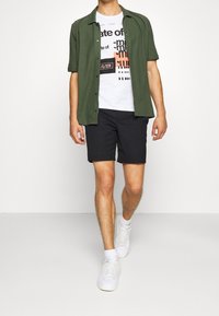 Zelená košile s krátkým rukávem přes bílý tričko s grafikou, spárovaná s černými šortkami a bílými teniskami. Oblečení má ležérní design.