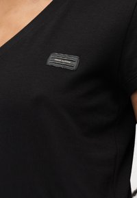 Επίκεντρο λεπτομέρειας μαύρου μπλουζιού με λαιμόκοψη V, με μικρό ορθογώνιο λάβαρο λογότυπου Armani Exchange στην περιοχή του άνω στήθους.