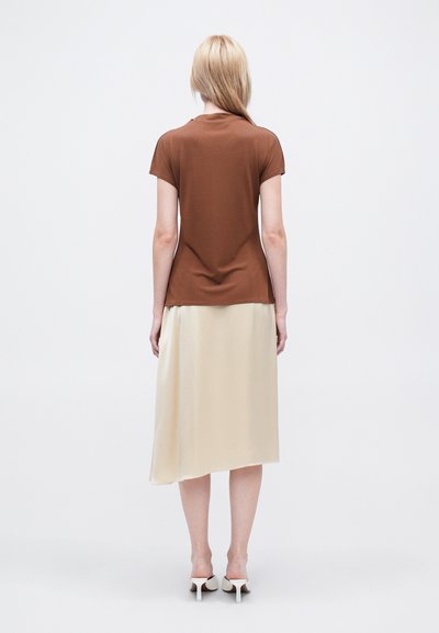 Tricou bronz cu mâneci scurte, asortat cu o fustă asimetrică de culoare crem. Fusta are o textură netedă și lucioasă.
