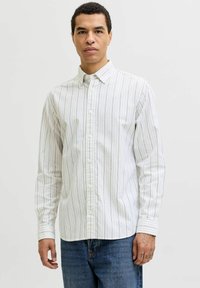 Homme portant une chemise blanche à boutons avec de fines rayures verticales et un jean bleu, debout devant un fond clair uni.