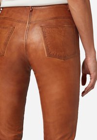 Braune Lederhosen mit glatter Textur, ausgestattet mit zwei Gesäßtaschen und einer taillierten Passform, die Schneider- und Nähdetails hervorhebt.