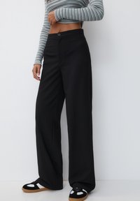 Wide-leg black trousers with a smooth texture, featuring a front button closure and crisp lines. Paired with black and white sneakers.