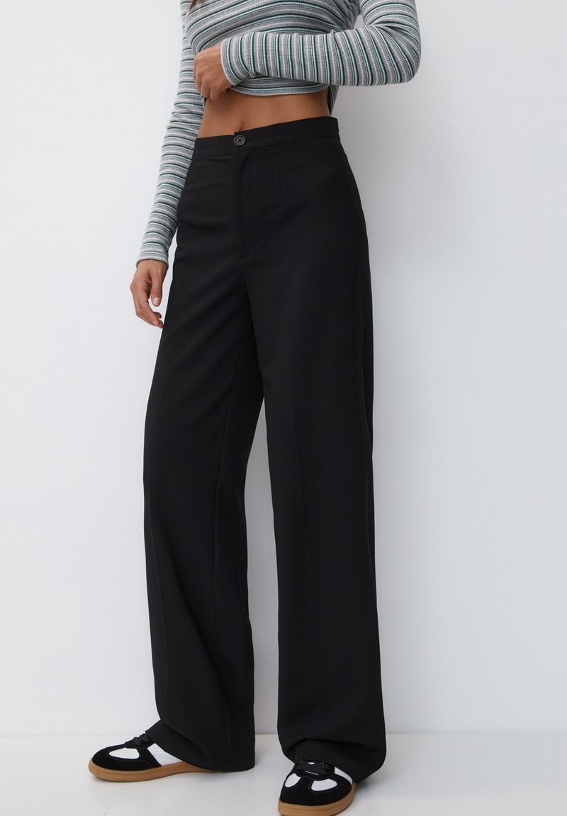 Wide-leg black trousers with a smooth texture, featuring a front button closure and crisp lines. Paired with black and white sneakers.