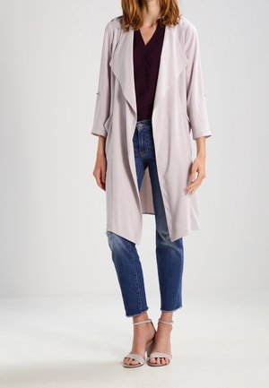 Frau trägt einen hellbeigen langen offenen Cardigan, ein dunkelviolettes Top, blaue kurze Jeans und beige Absatzschuhe mit Knöchelriemen und steht vor einem schlichten Hintergrund.