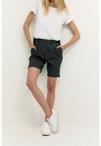 Femme portant un short noir à ourlet roulé, un t-shirt blanc et des baskets blanches, se tenant les mains dans les poches contre un fond blanc uni.